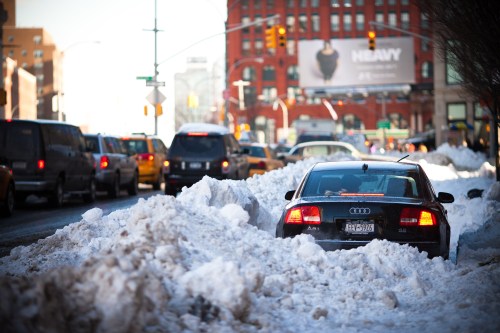 A black car stuck in deep snow on a busy city street, surrounded by snowbanks and traffic.