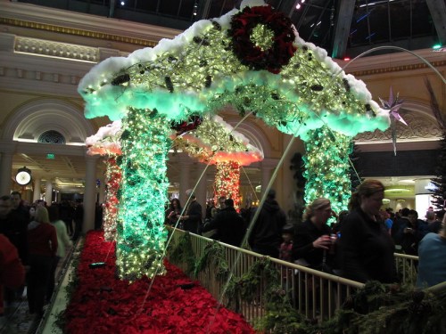 A festive display with illuminated arches and greenery, surrounded by crowds enjoying a holiday event indoors.
