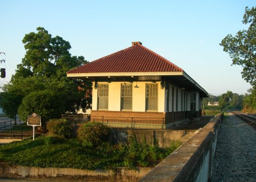 Historic train station with a red-tiled roof, surrounded by greenery and railway tracks under a clear blue sky.