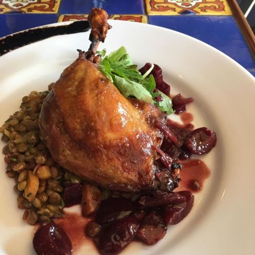 A plated dish featuring a crispy duck leg, lentils, greens, and a cherry sauce on a colorful table.