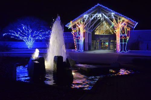 A brightly lit entrance with festive decorations, featuring a fountain and illuminated figures on either side.