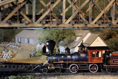 A model steam train passes under a wooden bridge, with a miniature scene of buildings and figures in the background.