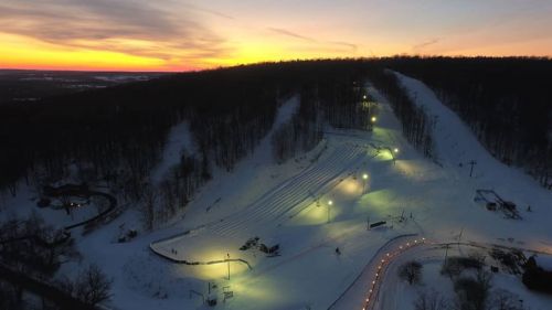 A snowy landscape at sunset, featuring ski slopes and illuminated paths winding through a wooded area.