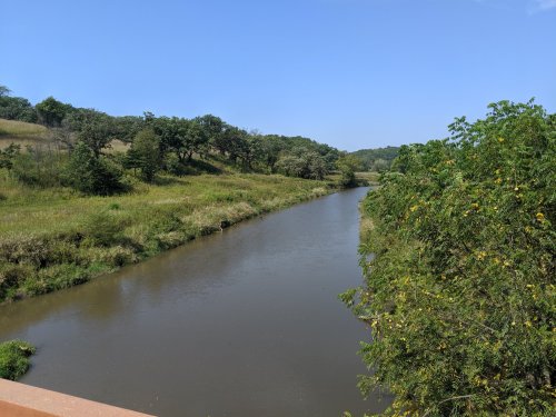 A serene river flows through lush green fields and trees under a clear blue sky.