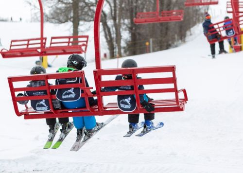 Three young skiers on a red chairlift, wearing helmets and ski gear, surrounded by snowy slopes and trees.