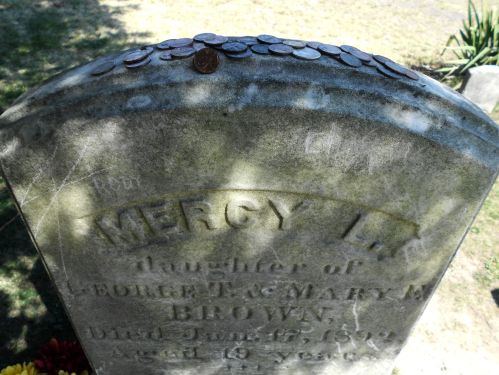 A weathered gravestone with the name "Mercy L." and coins placed on top, surrounded by grass and flowers.