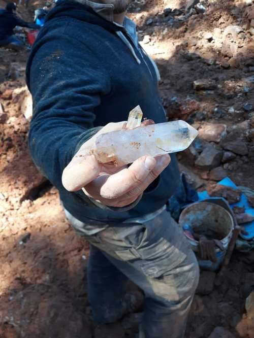 A person holds a large quartz crystal in a mining area, with dirt and tools visible in the background.