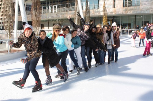 A group of people ice skating in a line, smiling and enjoying a sunny day at an outdoor rink.