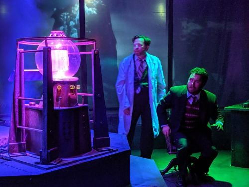 Two men in a lab setting, one in a lab coat and the other in a suit, with a glowing scientific apparatus.
