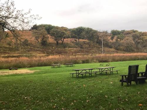 A grassy area with picnic tables and a backdrop of rolling hills and trees under a cloudy sky.