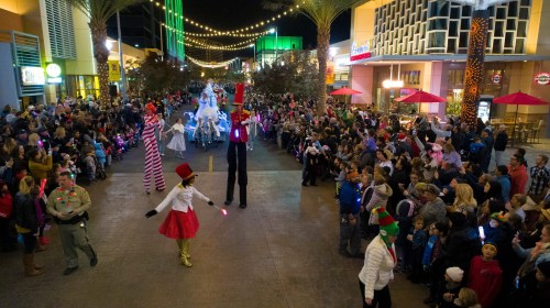 A festive parade with performers on stilts, colorful lights, and a crowd enjoying the celebration in a lively street.