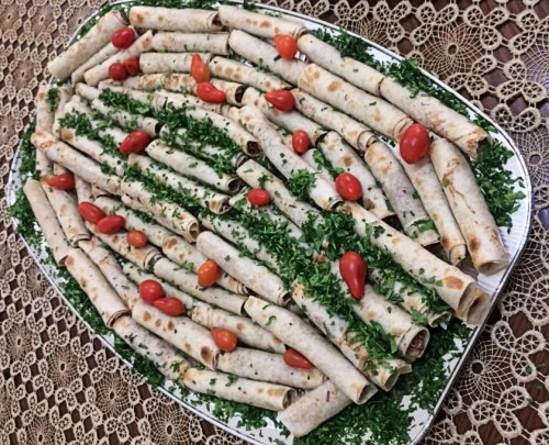 A platter of rolled flatbreads garnished with fresh parsley and cherry tomatoes, arranged neatly on a decorative tablecloth.