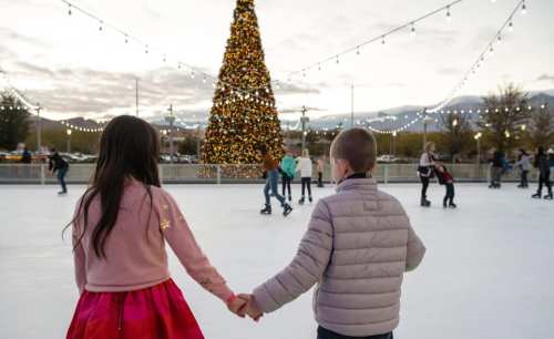 Two children hold hands while ice skating, with a large decorated Christmas tree in the background.