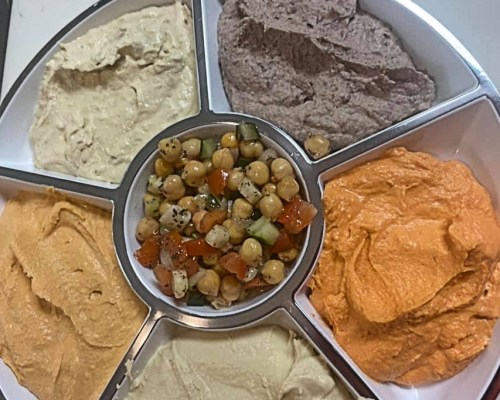 A platter with six varieties of hummus and a central salad of chickpeas, cucumbers, and diced vegetables.