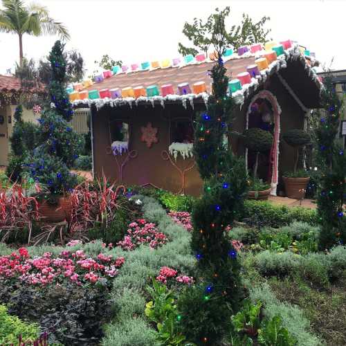 A whimsical gingerbread house surrounded by colorful flowers and festive lights in a lush garden setting.