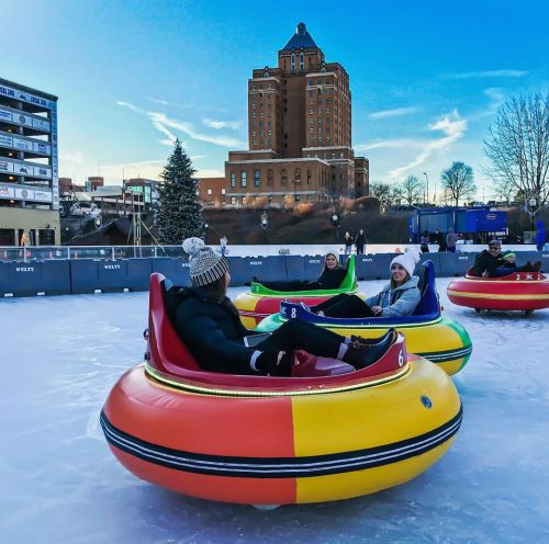 People enjoying a colorful bumper car ride on ice, with a historic building and clear blue sky in the background.