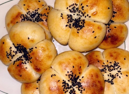 A plate of golden-brown, flower-shaped bread rolls topped with black sesame seeds.