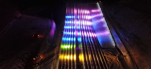 Aerial view of a brightly lit, colorful striped pattern on a snowy surface at night.