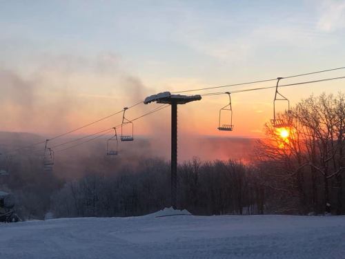 A ski lift stands against a colorful sunset, with snow-covered trees and mist in the background.