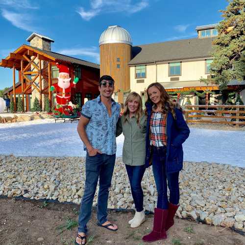 Three people pose together outdoors in front of a festive building, with holiday decorations and a snowy landscape.