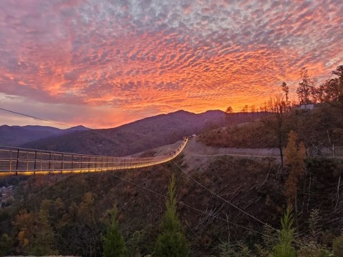 A suspension bridge stretches across a valley under a vibrant sunset with colorful clouds and mountains in the background.