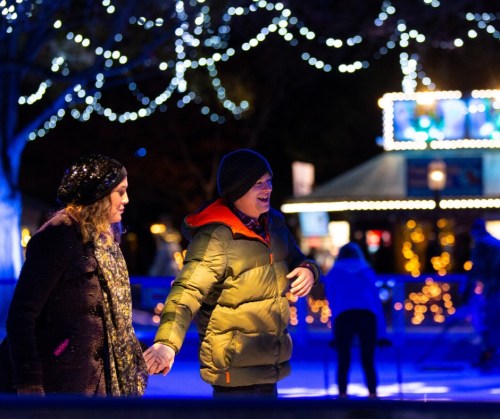 A couple ice skating together, holding hands, surrounded by festive lights and a winter atmosphere.