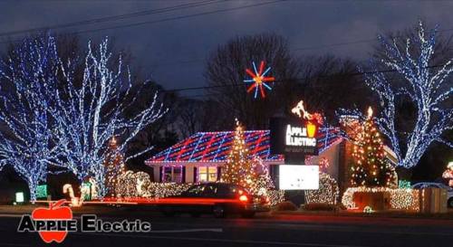A festive display of colorful holiday lights on a building and trees, with a sign for Apple Electric.