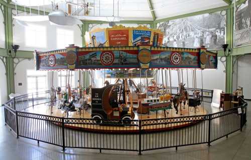 A vintage carousel with colorful decorations and wooden horses, enclosed by a black railing in a bright indoor space.