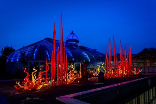 Colorful glass sculptures in red and orange stand outside a glass conservatory, illuminated against a twilight sky.