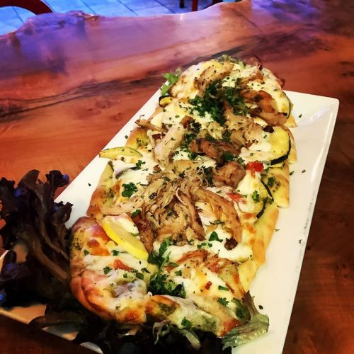 A platter of pizza topped with chicken, vegetables, and herbs, served on a wooden table with a side of greens.