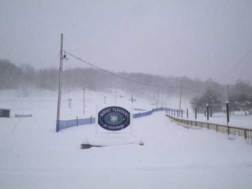 A snowy landscape with a sign for "Snowy Pleasant" partially buried in snow, surrounded by trees and ski lift poles.