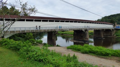 A covered bridge spans a river, with people walking on it and lush greenery surrounding the area.