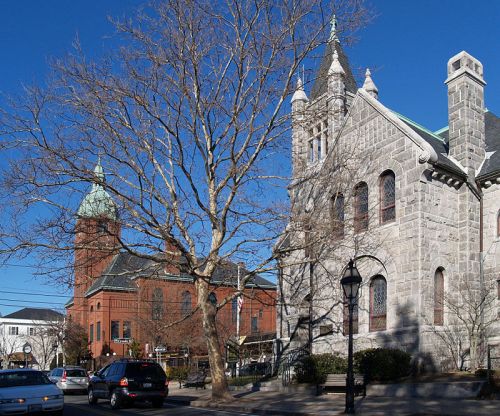 Historic stone and brick buildings with steeples, a bare tree, and a clear blue sky in a quaint town setting.