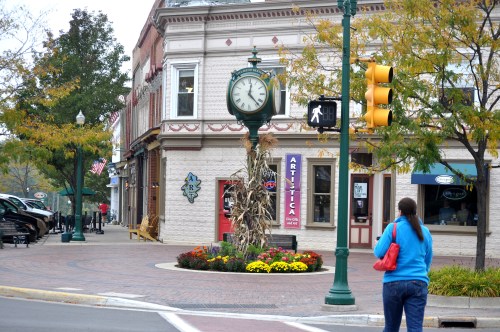 A street corner with a clock, traffic light, and shops, surrounded by colorful flowers and autumn trees.