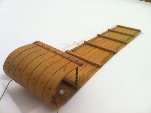 A wooden sled resting on a snowy surface, with a curved front and a long flat base.