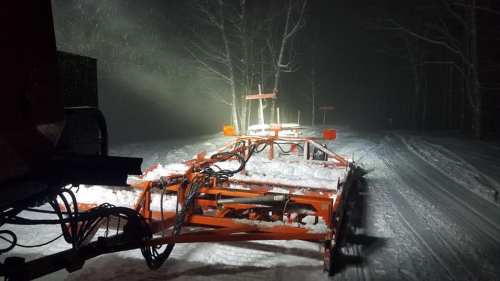 A snowplow clearing a snowy road at night, illuminated by headlights in a snowy, foggy environment.
