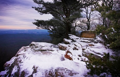 Snow-covered rocky outcrop with a tree, overlooking a vast mountainous landscape under a cloudy sky.