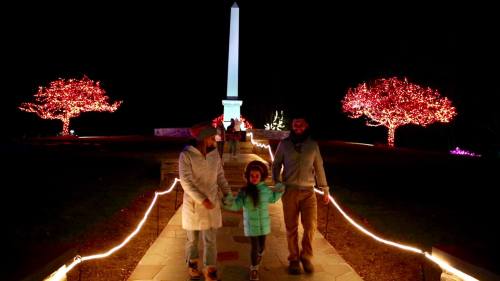A family walks together at night, surrounded by illuminated trees and a monument, creating a festive atmosphere.