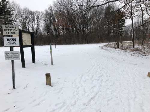 A snowy trail with a sign about riders and dogs, surrounded by bare trees in a winter landscape.