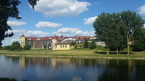 A scenic view of a riverside landscape featuring buildings, trees, and a blue sky with fluffy clouds.