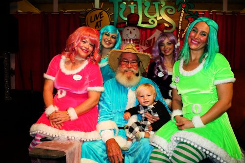 A group of six people in colorful costumes, including Santa, posing together in a festive setting.