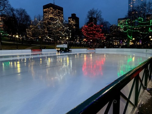 An ice skating rink at dusk, surrounded by trees adorned with colorful lights and city buildings in the background.