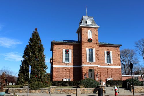 Historic brick building with a clock tower, decorated for the holidays, next to a large Christmas tree. Clear blue sky.