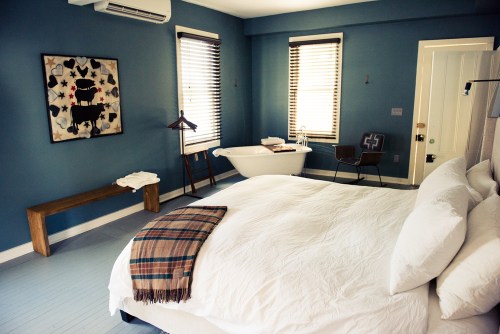A cozy bedroom with a white bed, blue walls, a vintage bathtub, and stylish decor, featuring natural light from windows.