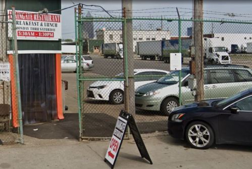 A restaurant entrance with a sign, surrounded by a fenced area and parked cars, in an industrial setting.