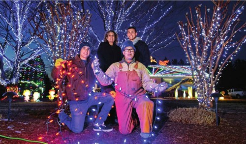 Four people pose with holiday lights in a festive outdoor setting, surrounded by illuminated trees at night.