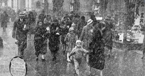 A snowy street scene with people, including children, walking in winter attire amidst heavy snowfall.