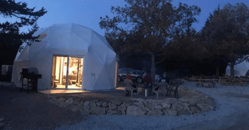 A geodesic dome structure at dusk, with a fire pit and chairs outside, surrounded by trees and gravel pathways.