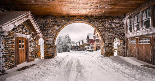 Snow-covered street framed by stone archway, with colorful houses and trees in a winter landscape.