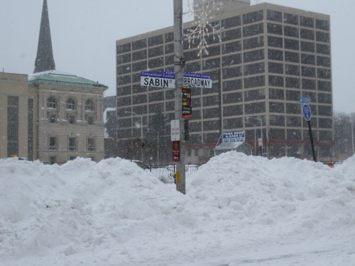 Snow-covered street corner with a sign for Sabin and Broadway, featuring buildings in the background and falling snow.